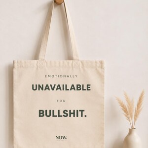 Könnte beinhalten: Eine beigefarbene Tragetasche aus Segeltuch mit zwei Schulterriemen. Auf der Vorderseite der Tasche befindet sich der schwarze Aufdruck: EMOTIONALLY UNAVAILABLE FOR BULLSHIT. Unter dem Text befindet sich ein kleines NDW-Logo. Die Tasche hängt an einem Holzhaken.