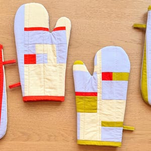 Może przedstawiać: Cztery pikowane rękawice kuchenne o różnych wzorach. Jedna ma czerwono-jasnoniebieskie pionowe paski, druga ma wzór w bloki w kolorze kremowym, jasnoniebieskim i czerwonym, trzecia ma wzór w bloki kremowo-jasnoniebiesko-oliwkowe, a czwarta ma oliwkowo-jasnoniebieskie paski.