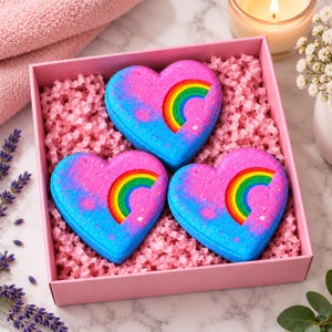 Puede incluir: Tres bombas de baño en forma de corazón dispuestas en una caja de regalo rosa rellena de papel arrugado rosa. Cada bomba de baño presenta una superficie con degradado azul y rosa vibrante con un arcoíris colorido en el centro.