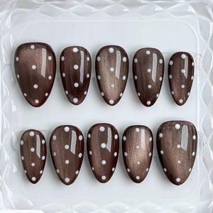 Puede incluir: Un juego de diez uñas postizas en forma de almendra con un acabado marrón oscuro metálico brillante y efecto ojo de gato. Cada uña está decorada con una fila vertical de pequeños puntos blancos espaciados uniformemente en el centro.