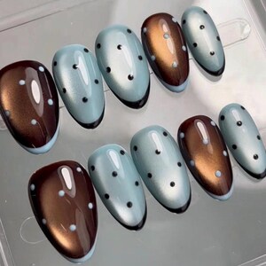 Puede incluir: Un juego de diez uñas postizas en forma de almendra con acabado brillante. El diseño alterna entre una base de color bronce metálico y una base azul claro brillante, ambas decoradas con pequeños puntos negros espaciados uniformemente.