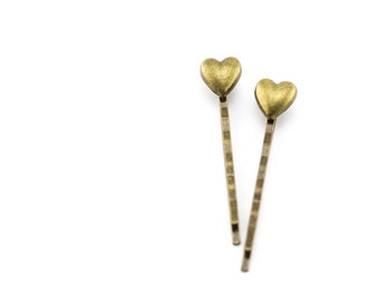 May include: Two antique gold-colored bobby pins. Each pin features a heart-shaped charm at the top. The pins are set against a white background, highlighting their design and color.