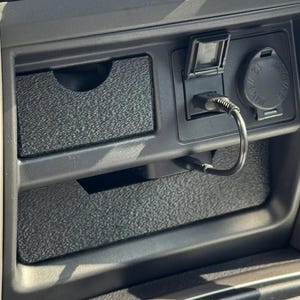 以下が含まれることがあります： テクスチャ仕上げの黒い自動車用センターコンソール収納インサート。USBケーブル用の専用スロットと、円形の電源コンセントカバーを備えています。このインサートは、既存のダッシュボードコンパートメントにフィットするように設計されています。