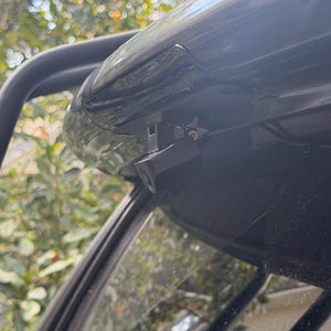 Puede incluir: Una pequeña cámara de visión trasera negra montada en el marco superior de una ventana trasera. La carcasa de la cámara es rectangular con una lente circular centrada en la parte frontal, unida a un soporte metálico en el exterior del vehículo.
