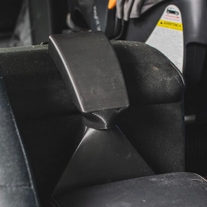 Pode incluir: Um suporte de plástico preto para encosto de cabeça de assento de carro com uma base triangular cónica e uma secção superior retangular. O suporte está fixado no topo de um encosto de cabeça de tecido preto entre dois apoios laterais.