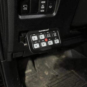 May include: A black Auxbeam switch panel mounted inside a vehicle dashboard. The rectangular device features eight illuminated buttons, with specific icons for backup and rear lights, and a central red power button, positioned below vehicle controls.