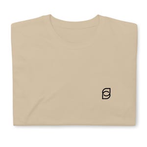 May include: A light tan crew neck t-shirt featuring a small, black, abstract geometric logo embroidered on the lower right side of the chest. The shirt is made of a solid-colored fabric and has a standard short-sleeve design with a ribbed collar.