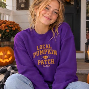 Könnte beinhalten: Ein violettes Sweatshirt mit Rundhalsausschnitt, das den Text "LOCAL PUMPKIN PATCH" in einer goldenen College-Schriftart zeigt, mit einer kleinen Kürbisgrafik in der Mitte und dem darunter gedruckten Text "Est. 1998", kombiniert mit einer hellblauen Jeans.
