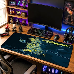 Op de afbeelding: Een rechthoekige bureaumat met een donkerblauwe achtergrond, een gele kaart van Night City en het Cyberpunk 2077-logo in gele tekst. De mat ligt op een houten bureau voor een computermonitor en een toetsenbord.