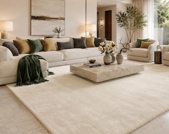 Puede incluir: Una alfombra grande y rectangular de color crema con una textura suave y afelpada que cubre el suelo de una sala de estar. La alfombra presenta un tono sólido y neutro que complementa los muebles circundantes, incluyendo un sofá y una mesa.