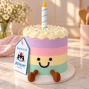 May include: A plush toy shaped like a multi-coloured layer cake with pastel purple, pink, yellow, and green stripes. It features a smiling face, two brown legs, a white candle on top, and a tag that reads Gracie is 11 and JELLYCAT LONDON.