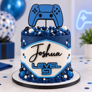 May include: A cylindrical cake decorated in shades of blue, white, and black. The cake features a blue gaming controller topper, a central white panel with the name Joshua and the text Level 9, and blue frosting swirls with small round sprinkles.