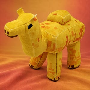 May include: A plush camel toy featuring a blocky, pixelated design in shades of yellow and orange. The stuffed animal has a rectangular body, a single hump on its back, four sturdy legs, and a stylized head with dark brown eyes and nostrils.