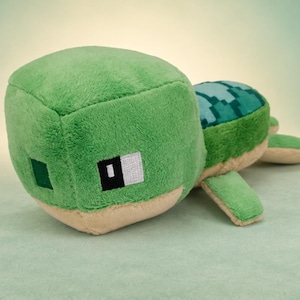 Peut inclure: Une peluche en forme de tortue au design cubique et pixélisé, avec une tête et un corps vert vif, un dessous beige et une carapace vert foncé à motifs. Le visage présente des yeux carrés noirs et blancs et un petit carré vert sur le côté.
