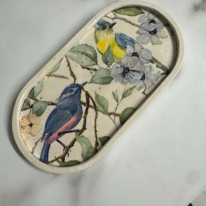 Op de afbeelding: Een ovaalvormig, gebroken wit dienblad met een decoratieve print van twee vogels op takken met groene bladeren en lichtpaarse bloemen. De ene vogel is blauw en geel, terwijl de andere voornamelijk blauw is met een rozeachtige borst.