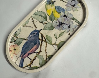 May include: An oval-shaped off-white tray featuring a decorative print of two birds perched on branches with green leaves and light purple flowers. One bird is blue and yellow, while the other is primarily blue with a pinkish-toned chest area.