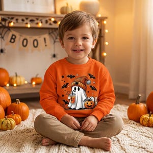 Puede incluir: Una sudadera de manga larga de color naranja brillante con un estampado gráfico de un fantasma blanco con un gorro a rayas, sosteniendo un café helado y rodeado de murciélagos negros, estrellas y una pequeña calabaza en el centro.