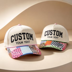 May include: Two off-white baseball caps featuring the black embroidered text CUSTOM YOUR TEXT on the front panels. The brims display a colorful patchwork pattern with pink, blue, and green geometric shapes, stripes, and polka dots.