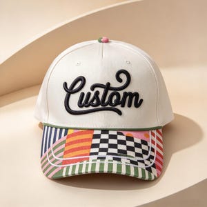 May include: A cream-colored baseball cap featuring the word Custom embroidered in black script on the front panel. The brim displays a multi-colored patchwork pattern with stripes, checks, and dots, accented by a green cord trim.