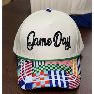 May include: A cream-colored baseball cap featuring the embroidered black text Game Day on the front panel. The cap includes a green cord trim above the brim, which is decorated with a vibrant, multi-colored patchwork pattern of checks, stripes, and dots.