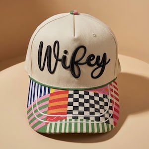 May include: A cream-colored baseball cap featuring the word Wifey embroidered in black script across the front. The brim displays a multi-colored patchwork pattern including black and white checkers, vertical stripes in green, pink, and blue, and orange blocks.