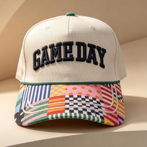 May include: A cream-colored baseball cap featuring the text GAME DAY embroidered in dark navy blue letters across the front. The cap includes a green cord trim above the brim, which is decorated with a colorful patchwork pattern of stripes, dots, and checks.