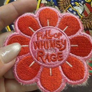 May include: A circular embroidered patch shaped like a flower with orange petals and a pink center. The center features the embroidered text "FULL OF WHIMSY AND RAGE" in orange thread. The patch has a white stitched border and a textured fabric finish.