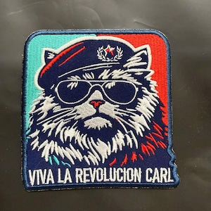 May include: A rectangular embroidered patch featuring a cat wearing a dark blue beret with a red star emblem and black sunglasses. The background is split into turquoise and red sections. The text VIVA LA REVOLUCION CARL is in white.