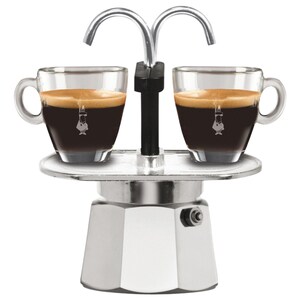 May include: A silver metal espresso maker with a dual-spout top, featuring two clear glass espresso cups filled with dark coffee and topped with crema. Each glass cup displays a small white logo of a person with a moustache on the front.