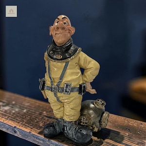 Op de afbeelding: Een beeldje van een kaal personage in een geel vintage diepzeeduikpak met een zwarte kraag, donkergrijze laarzen en een riem met rechthoekige zakjes. Een metallic bronskleurige duikhelm staat op de grond naast het figuurtje.
