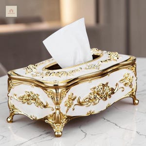 Puede incluir: Un soporte rectangular para caja de pañuelos con base blanca y detalles florales en relieve de color dorado. La caja se apoya sobre cuatro patas curvas doradas e incluye un pañuelo blanco que sobresale por la abertura central.