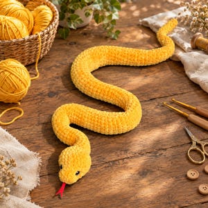 Puede incluir: Una serpiente larga de ganchillo en color amarillo, suave y afelpada, con una lengua de fieltro roja. La serpiente tiene una superficie texturizada y está colocada en forma sinuosa. Presenta pequeños ojos negros y lana gruesa y esponjosa.