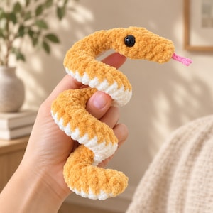 May include: A coiled plush snake toy crafted from soft, fuzzy yarn in alternating bands of golden yellow and white. The snake features a smooth, round black eye and a small, protruding pink tongue made of yarn, creating a tactile, textured appearance.