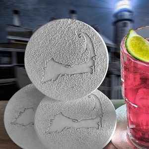 May include: Four gray coasters with a raised design of the outline of Cape Cod. The coasters are stacked on top of each other. A glass of pink lemonade with a lime wedge is sitting on a coaster.