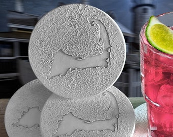 May include: Four gray coasters with a raised design of the outline of Cape Cod. The coasters are stacked on top of each other. A glass of pink lemonade with a lime wedge is sitting on a coaster.