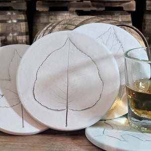 May include: Four white ceramic coasters with leaf designs. The coasters are arranged in a semi-circle with a glass of amber liquid in the center.
