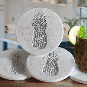 May include: Four white ceramic coasters with a pineapple design. The coasters are round and have a textured surface. A copper mug with a drink and a cucumber slice is sitting on top of the coasters.