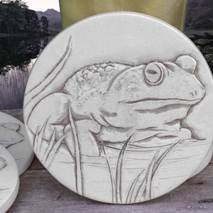 May include: A white ceramic coaster with a detailed embossed design of a frog sitting in a pond. The frog is facing the right side of the coaster and has a textured, realistic appearance.