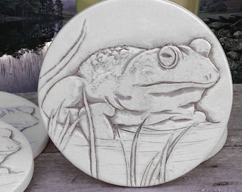 May include: A white ceramic coaster with a detailed embossed design of a frog sitting in a pond. The frog is facing the right side of the coaster and has a textured, realistic appearance.