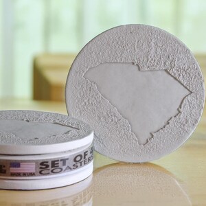 May include: Set of four white ceramic coasters featuring the outline of the state of South Carolina. The coasters are stacked and have a label that reads "Set of 4 Coasters" and "Made in USA".