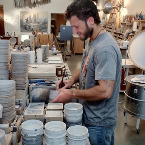 May include: A man in a grey t-shirt and blue jeans is working on a project in a pottery studio. He is surrounded by ceramic pieces, including a stack of round plates and several white buckets. The studio is well-lit and has a variety of tools and equipment.