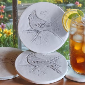 May include: Set of three white ceramic coasters with an embossed design of a cardinal bird perched on a branch. The coasters are stacked on top of each other.