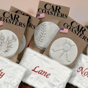May include: Four white ceramic coasters with different designs, including a deer, a baseball, a golf ball and tee, and a bicycle. The coasters are packaged in brown paper bags with the text "CAR COASTERS" printed on them. The coasters are displayed in white stockings with the names "Andy", "Lane", and "Noah" embroidered on them.