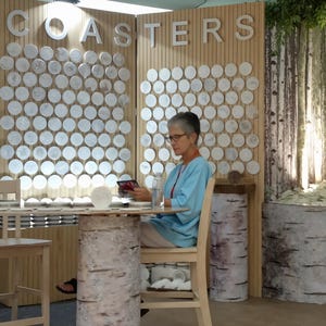 May include: A woman sits at a table in a shop, looking at a phone. The table is made of wood and has a birch bark design. The walls are decorated with a display of round white coasters with various designs. The sign above the coasters reads "COASTERS".