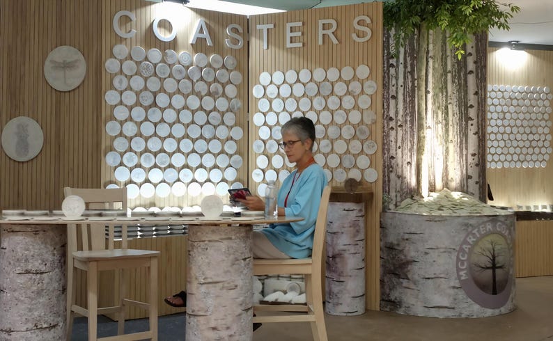 May include: A woman sits at a table in a shop, looking at a phone. The table is made of wood and has a birch bark design. The walls are decorated with a display of round white coasters with various designs. The sign above the coasters reads "COASTERS".