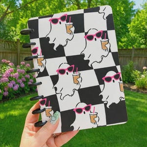 May include: A disc-bound notebook featuring a repeating pattern of white ghosts wearing bright pink sunglasses and holding tan iced coffee cups against a black and white checkered background. The notebook has black plastic binding discs along the side.