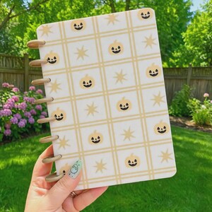 Op de afbeelding: Een notitieboek met schijfbinding en een witte kaft met een lichtbruin ruitpatroon. Het ontwerp bevat herhalende kleine lichtoranje pompoenen met zwarte gezichten en lichtbruine sterren die over het raster verspreid staan.