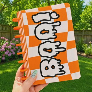 May include: A rectangular notebook with a bright orange and white checkered pattern on the cover. The word BOO is printed in large, black-outlined, white, dripping-style letters. The notebook features orange plastic binding discs on the side.