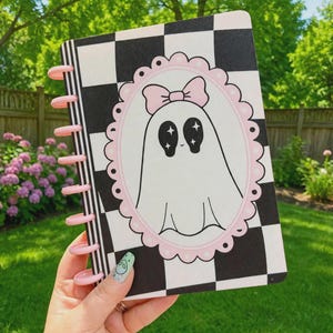 May include: A disc-bound notebook featuring a black and white checkered pattern cover. Centered on the cover is a pink scalloped oval frame containing a white ghost illustration with a pink bow and black star-filled eyes. The binding discs are pink.