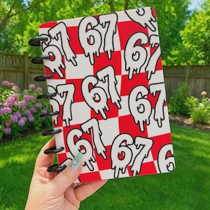 May include: A rectangular notebook with a red and white checkered pattern featuring a repeating black-outlined number 67 design with a dripping effect. The notebook is bound with black plastic discs along the left side and has a smooth cover.